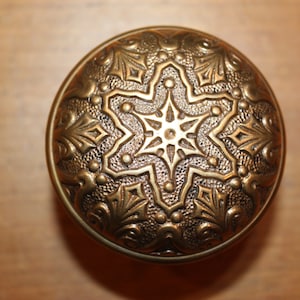 May include: A detailed, antique brass knob with an ornate, starburst design. The knob features a central eight-pointed star surrounded by floral and geometric patterns. The metal has a warm, aged patina.