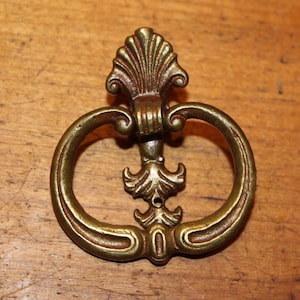 May include: Ornate brass drawer pull with a ring handle and decorative floral accents. The hardware has a vintage aesthetic and is set against a wooden surface.