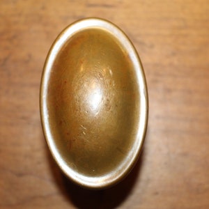 May include: Oval-shaped, antique brass-colored hardware. The hardware has a slightly raised, rounded center and a contrasting white rim. The object is set against a wooden background.