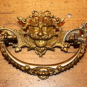 May include: Ornate brass drawer pull with a central grotesque mask design. The pull features a curved handle and decorative flourishes, suggesting a vintage or antique style. The brass has a warm, aged patina.