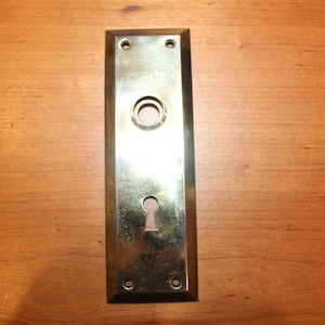May include: A brass door plate with a keyhole and a round hole for a doorknob. The plate has a rectangular shape with rounded corners.