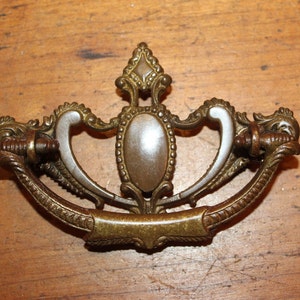 May include: A vintage brass drawer pull with a decorative design. The pull has a silver-toned center with a beaded edge and two ornate side pieces.