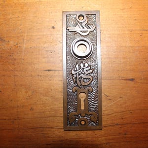 May include: A vintage brass door plate with an ornate design and a keyhole. The plate has a textured surface and a circular knob.