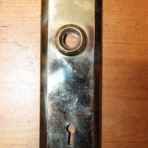 May include: A brass door plate with a keyhole and a round hole for a knob. The plate has a rectangular shape with rounded corners and is slightly tarnished.