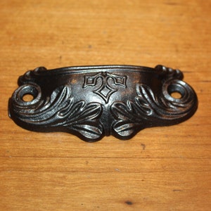 May include: Ornate black metal drawer pull with a decorative design. The handle features a central emblem and scrolling leaf patterns on each end. Perfect for adding a vintage touch to furniture.