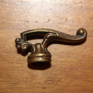 May include: A brass decorative hardware piece with a curved handle and ornate details. The handle is shaped like a stylized bird's wing.