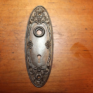May include: An antique bronze-colored door plate with an oval shape. The plate features ornate floral and scrollwork designs around the edges, with a keyhole and a circular opening for a doorknob.