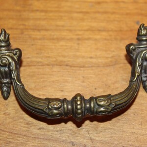 May include: A vintage brass drawer pull with ornate details and a flame-like design on each end.