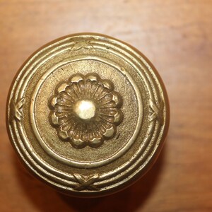 May include: A gold-toned metal knob with a decorative floral design in the center. The knob has a circular shape with concentric rings around the center.