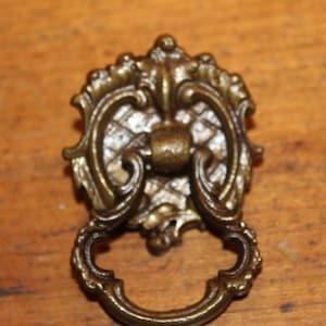 May include: A vintage ornate brass drawer pull with a decorative, intricate design. The pull has a ring handle and a detailed, scroll-like pattern.