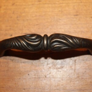 May include: A bronze-colored cabinet pull with a decorative, ornate design. The pull has a curved shape with detailed leaf-like patterns on each end and a central, rounded section. The hardware is likely for furniture or cabinetry.