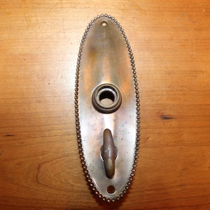 May include: An oval-shaped brass door plate with a beaded edge. The plate has a keyhole and a hook.