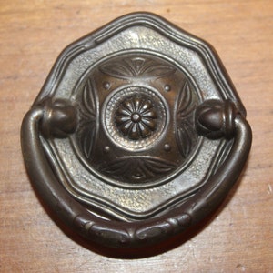 May include: A vintage octagonal-shaped cabinet pull with a decorative floral design and a ring handle. The pull is made of metal and has a dark bronze finish.