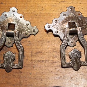 May include: Two antique bronze drawer pulls. Each pull features a decorative backplate with a textured pattern and a detailed handle. The pulls are mounted on a wooden surface, showcasing their vintage design and craftsmanship.