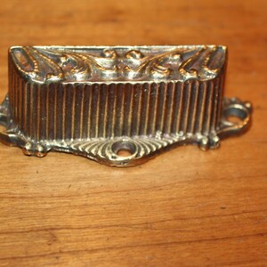 May include: A gold-toned metal drawer pull with a decorative, ornate design. The pull has a ribbed center section and a scalloped edge.