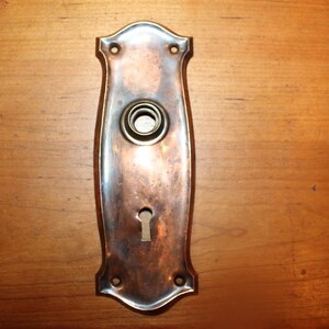 May include: A vintage brass door plate with a keyhole and a round hole for a doorbell. The plate has a slightly curved shape and a worn, aged finish.
