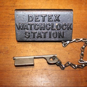 May include: A vintage DETEX WATCHCLOCK STATION key and station plate. The plate is dark gray with raised lettering. The key is silver-colored and attached to the plate with a chain. The items are on a wooden surface.