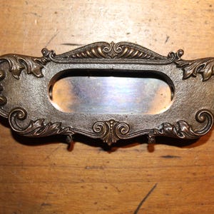 May include: Ornate bronze-colored drawer pull with an oval recessed area. The decorative hardware features scrolling leaf and floral designs. The drawer pull is likely vintage or antique, and is set against a wooden surface.
