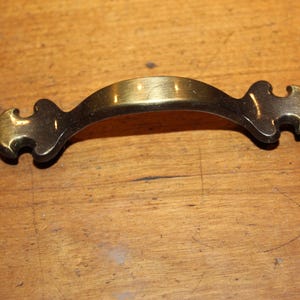 May include: A brass cabinet pull with a decorative, ornate design. The pull has a curved shape and a slightly tarnished finish.