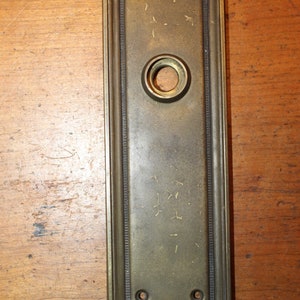 May include: A brass door plate with a keyhole and two small holes. The plate has a decorative border and is slightly tarnished.