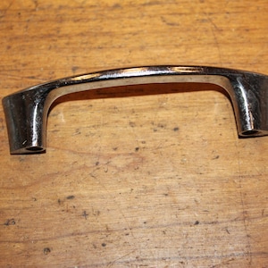 May include: A chrome-finished cabinet pull handle with a curved design. The handle has a rectangular shape with rounded edges and two mounting holes. The handle is set against a wooden surface.