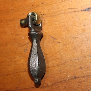 May include: A vintage, dark metal drawer pull with a brass roller and a teardrop-shaped handle. The hardware is set against a warm, wooden surface, showcasing its antique design and potential for furniture restoration projects.