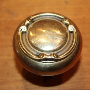 May include: A round, brass door knob with an ornate design. The knob has a smooth, polished finish and a raised, circular center.