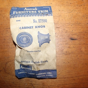 May include: A white card with blue text and illustrations of two cabinet knobs. The card reads "Amerock Furniture Trim", "No. ET590", "Cabinet Knob", "Antique English Finish", "American Cabinet Hardware Corp.", and "Rockford, Illinois".