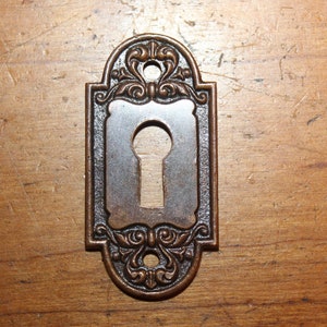 May include: A decorative keyhole cover with an ornate design in a bronze finish. The cover has a rectangular shape with a keyhole in the center.