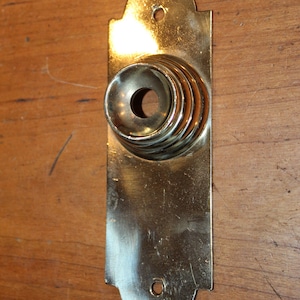 May include: A gold-toned doorbell button with a decorative, layered design. The button is mounted on a wooden surface.