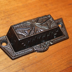 May include: A dark bronze-colored drawer pull with an ornate, rectangular handle and decorative backplates. The handle features a detailed floral and geometric pattern. The backplates have a diamond pattern and circular holes for mounting.