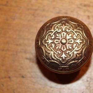 May include: Ornate, antique brass door knob with a detailed floral design. The knob is round with a slightly domed top and is set against a warm, wooden background. The metal has a rich, aged patina.
