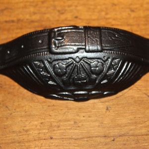 May include: Black cast iron drawer pull with a decorative belt buckle design. The handle features an ornate floral pattern and two mounting holes on either end. This vintage-style hardware is suitable for cabinets and furniture.