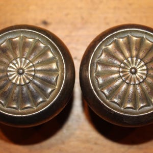 May include: Two antique bronze door knobs. Each knob has a circular design with a central rosette and radiating petal-like details. The knobs have a dark, aged patina, suggesting vintage or historical use. The background is a wooden surface.