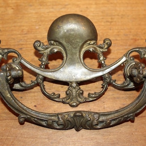 May include: Antique brass drawer pull with ornate scrollwork and a rounded top. The pull has a decorative design and is in good condition.