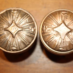 May include: Two antique bronze-colored cabinet knobs. Each knob features a central diamond shape with radiating lines, and a circular border. The knobs are likely made of metal and are designed for furniture.