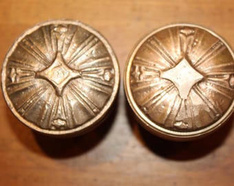 May include: Two antique bronze-colored cabinet knobs. Each knob features a central diamond shape with radiating lines, and a circular border. The knobs are likely made of metal and are designed for furniture.