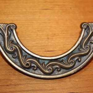 May include: A decorative, ornate, brass-colored, half-circle pull handle with a scroll design.