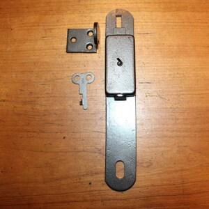 May include: A silver metal latch with a keyhole and a matching key. The latch has a rectangular plate with two holes and a round knob. The key is a simple, silver metal key with two loops.