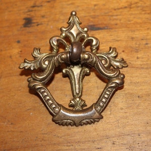 May include: Ornate brass drawer pull with a decorative design. The pull features a central ring and an elaborate, symmetrical pattern with floral and scroll elements. The hardware is set against a wooden surface.