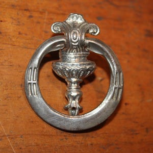 May include: A silver-toned drawer pull with an ornate design. The pull features a circular handle with decorative elements and a central, detailed fixture. The hardware is set against a warm, wooden surface.