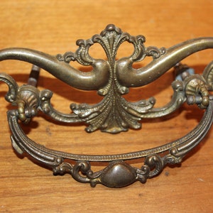 May include: Ornate brass drawer pull with a decorative, floral design. The pull has a curved shape and is attached to a wooden surface.