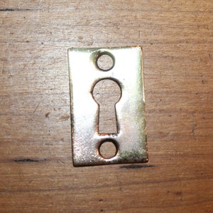 May include: A brass keyhole cover with a rectangular shape and a keyhole cut out in the center.