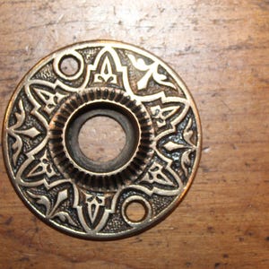 May include: Ornate, antique brass door plate with a central hole and decorative floral and geometric patterns. The circular plate has two smaller holes and is set against a wooden surface.