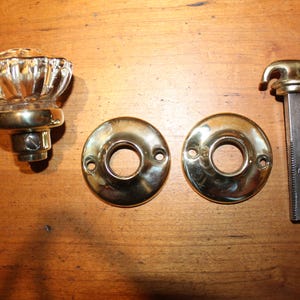 May include: An assortment of antique door hardware, including a faceted glass and brass doorknob, two brass backplates, and a brass and steel spindle. The hardware is arranged on a wooden surface.