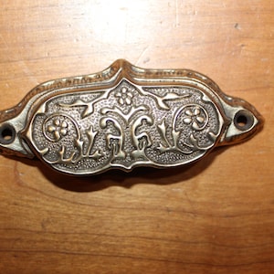 May include: A brass drawer pull with an ornate floral design. The pull is oval shaped and has two holes for mounting.