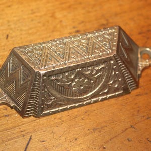 May include: A metallic drawer pull with a trapezoidal shape and a brass-toned finish. The surface features intricate embossed patterns, including geometric zig-zag lines on the sides and a central semi-circular floral motif with screw holes.