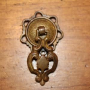 May include: An antique brass drawer pull with an ornate design. The pull features a circular backplate with a decorative edge, a central handle, and a stylized teardrop-shaped pull. The hardware is a warm, aged brass color.