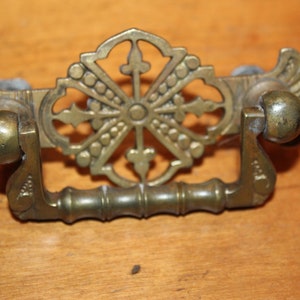 May include: A brass drawer pull with a decorative, ornate, circular design in the center and two rounded knobs on either side.