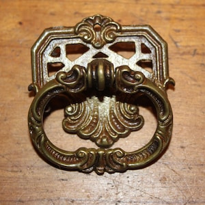 May include: Antique brass cabinet pull with a decorative, ornate design. The pull features a ring handle with intricate scrollwork and a decorative backplate with a geometric pattern.